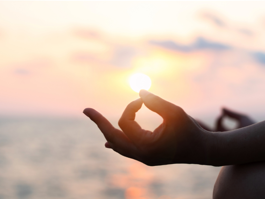 A hand is centered, the ocean andsun in the background. The hand's thumb and pointer finger are softly head together, forming an O.